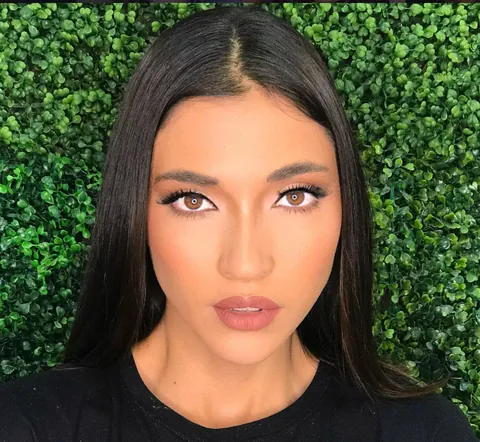 Cabelo liso escuro, olhar iluminado e boca nude diante de uma parede de folhagem